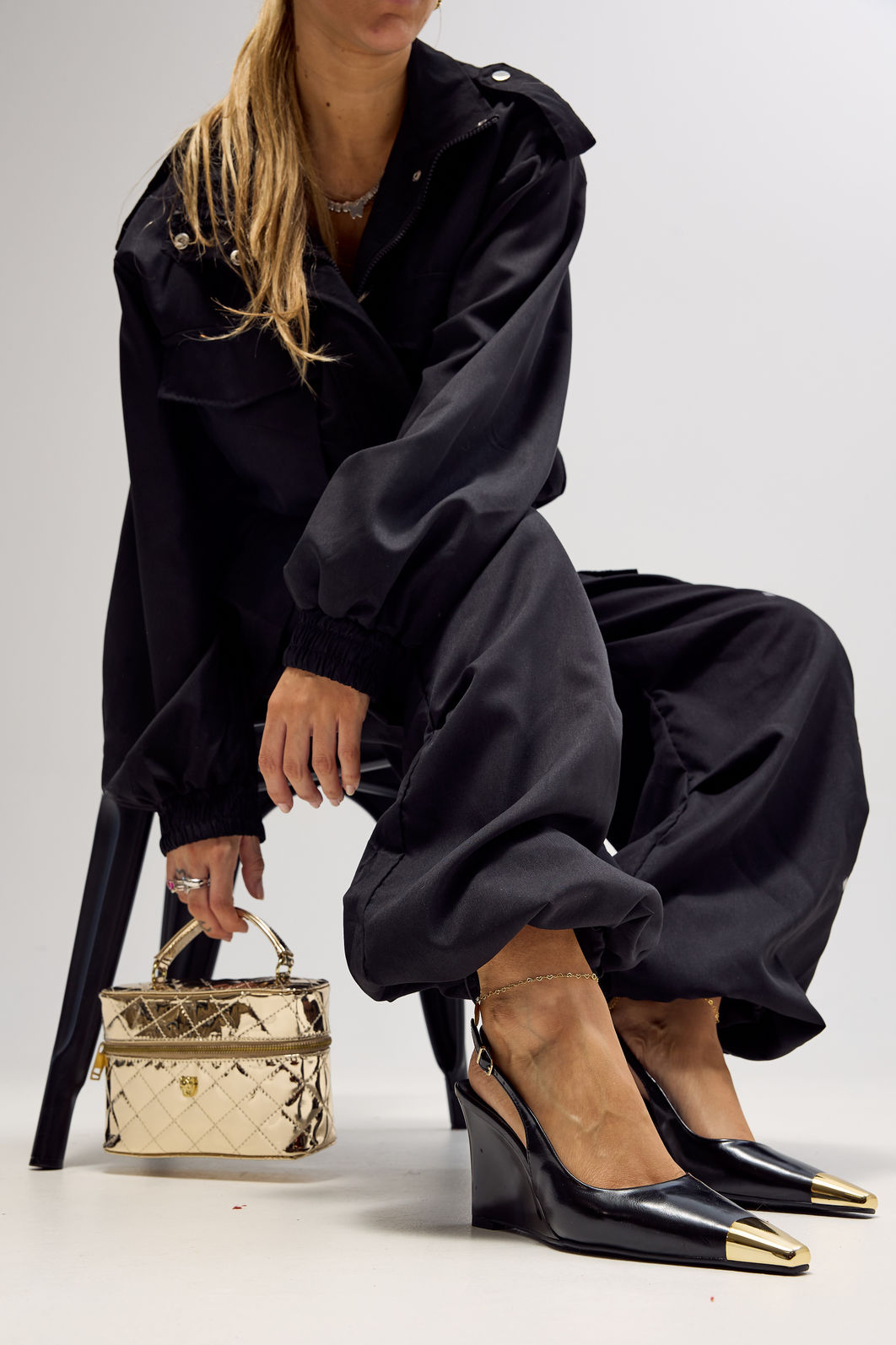 Slingback Naomi Preto Editorial Modelo segurando bolsa metalizada dourada enquanto usa slingback feminino Naomi preto com salto anabela e bico metálico dourado em destaque.