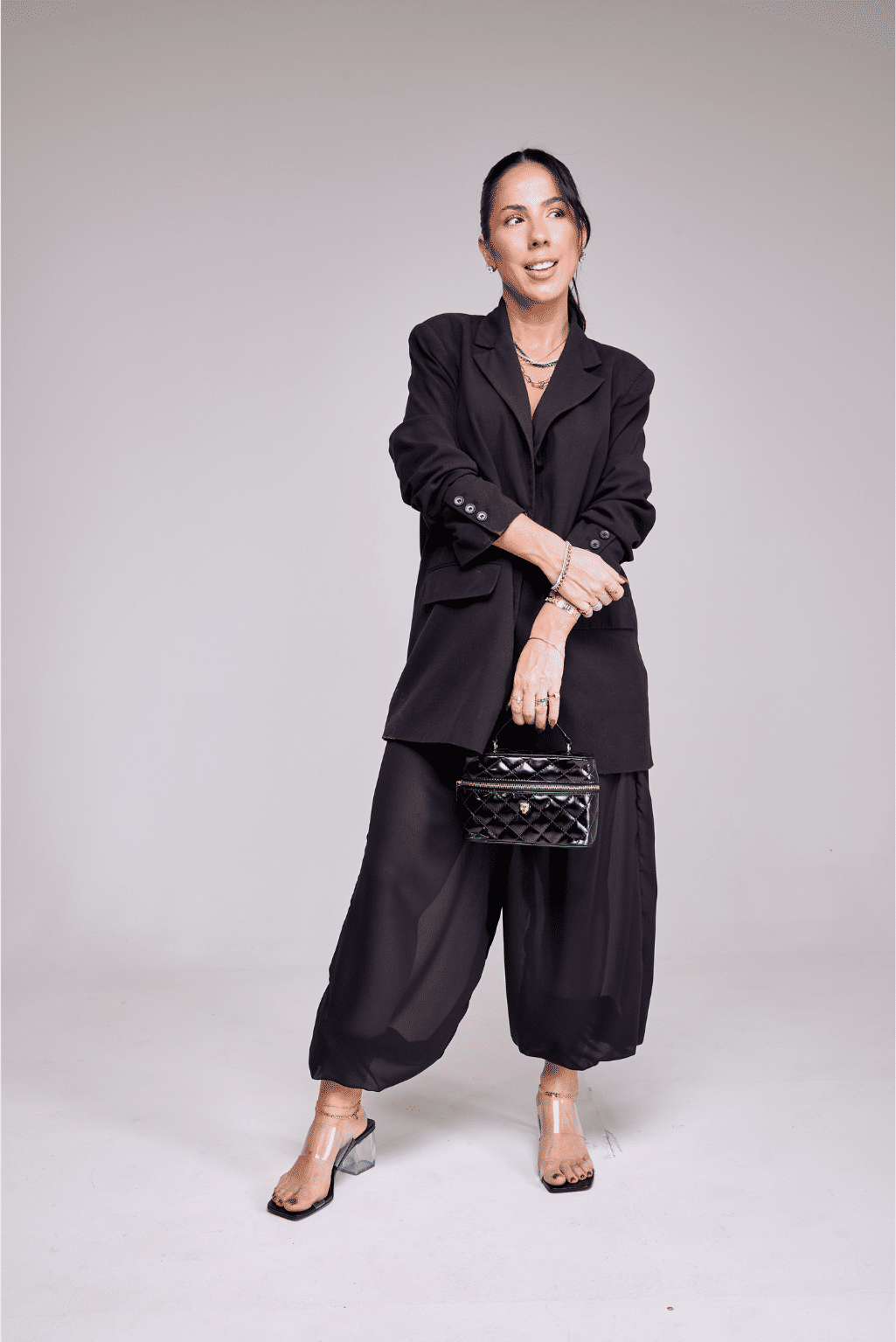 tamanco-feminino-salto-transparente-tiras-de-vinil-preto-kim Modelo usando look total black com blazer e bolsa preta, calçando o Kim - Tamanco Feminino Salto Transparente Tiras de Vinil Preto, corpo inteiro