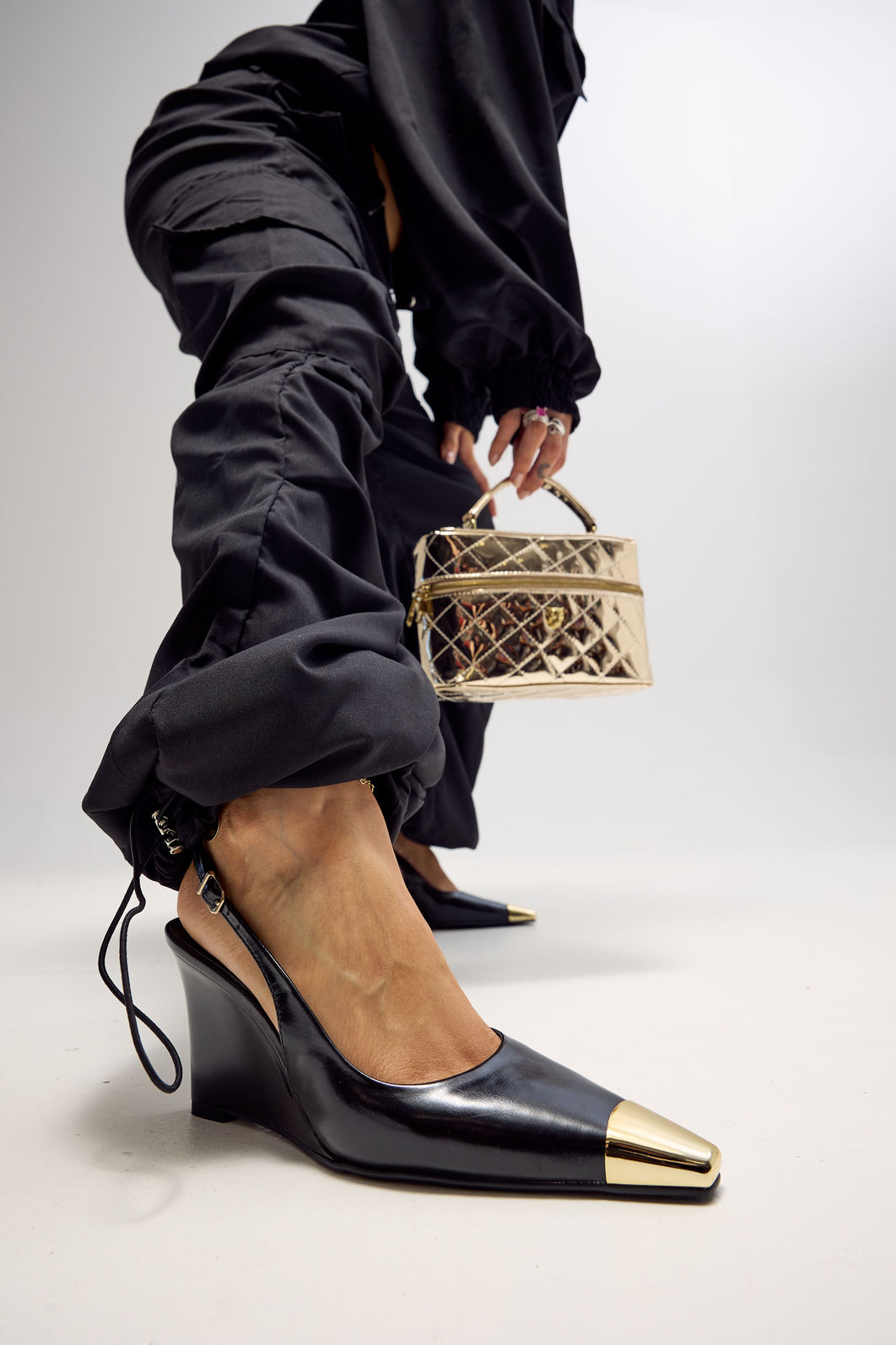 Close Slingback Naomi Preto Close da modelo usando slingback feminino Naomi preto com salto anabela de 9 cm e bico metálico dourado, acompanhado de bolsa metalizada dourada.