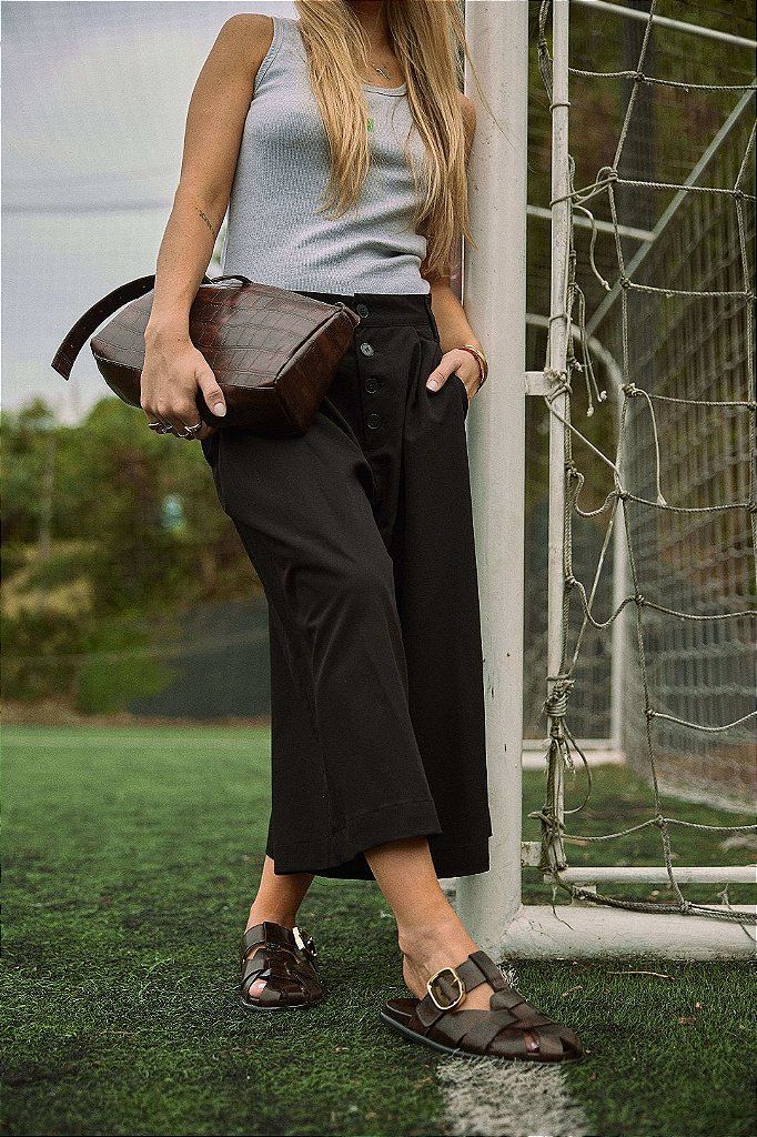 clog-feminino-fivela-marrom-aura Modelo de corpo inteiro usando o clog marrom com fivela dourada, calça pantalona preta e bolsa de couro croco, encostada na trave de um campo de futebol.