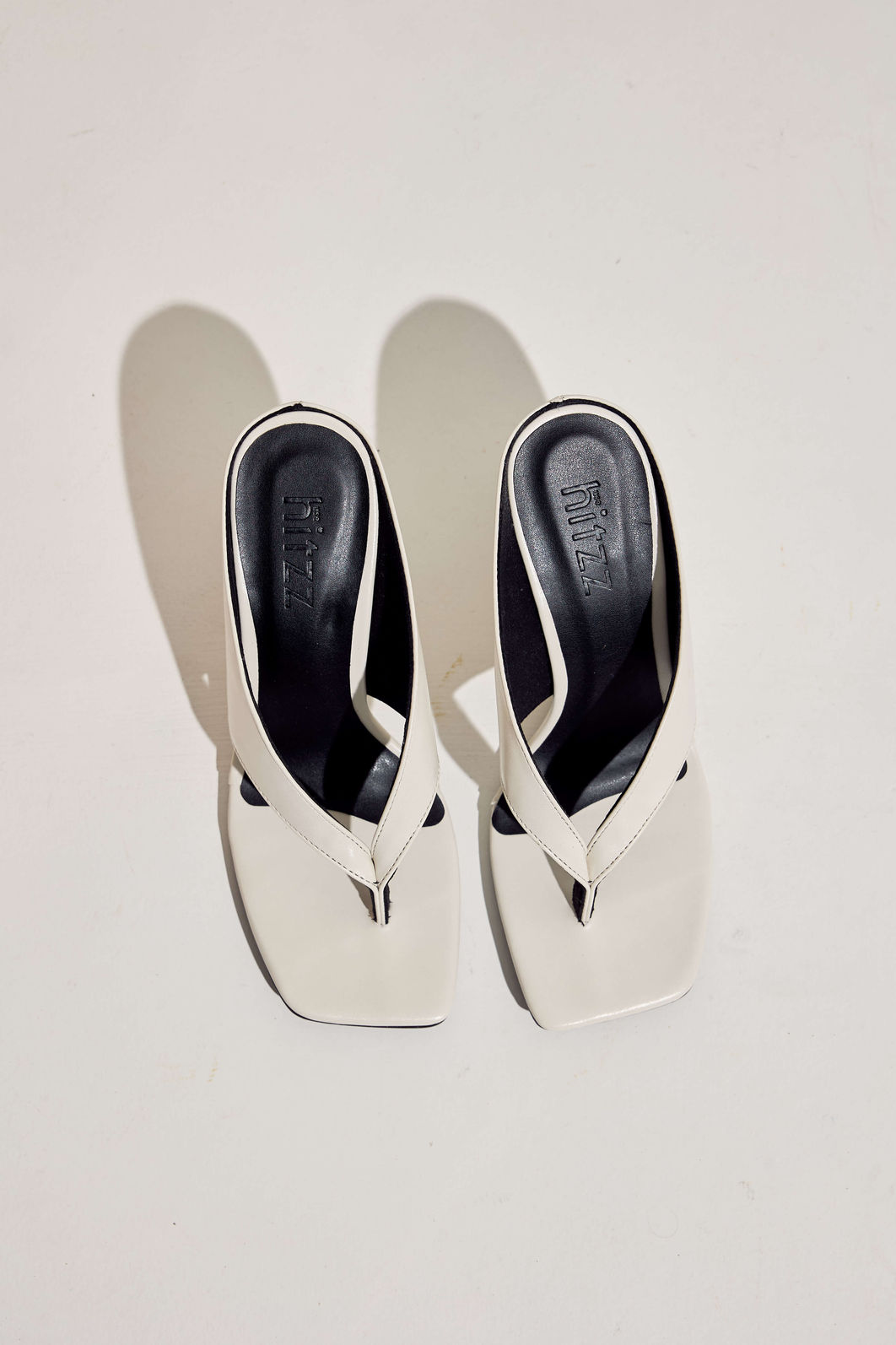 tamanco-feminino-dedo-salto-anabela-off-white-misaki Par de tamancos dedo off white vistos de cima, palmilha preta com a marca hitzz gravada, sombra em fundo claro.