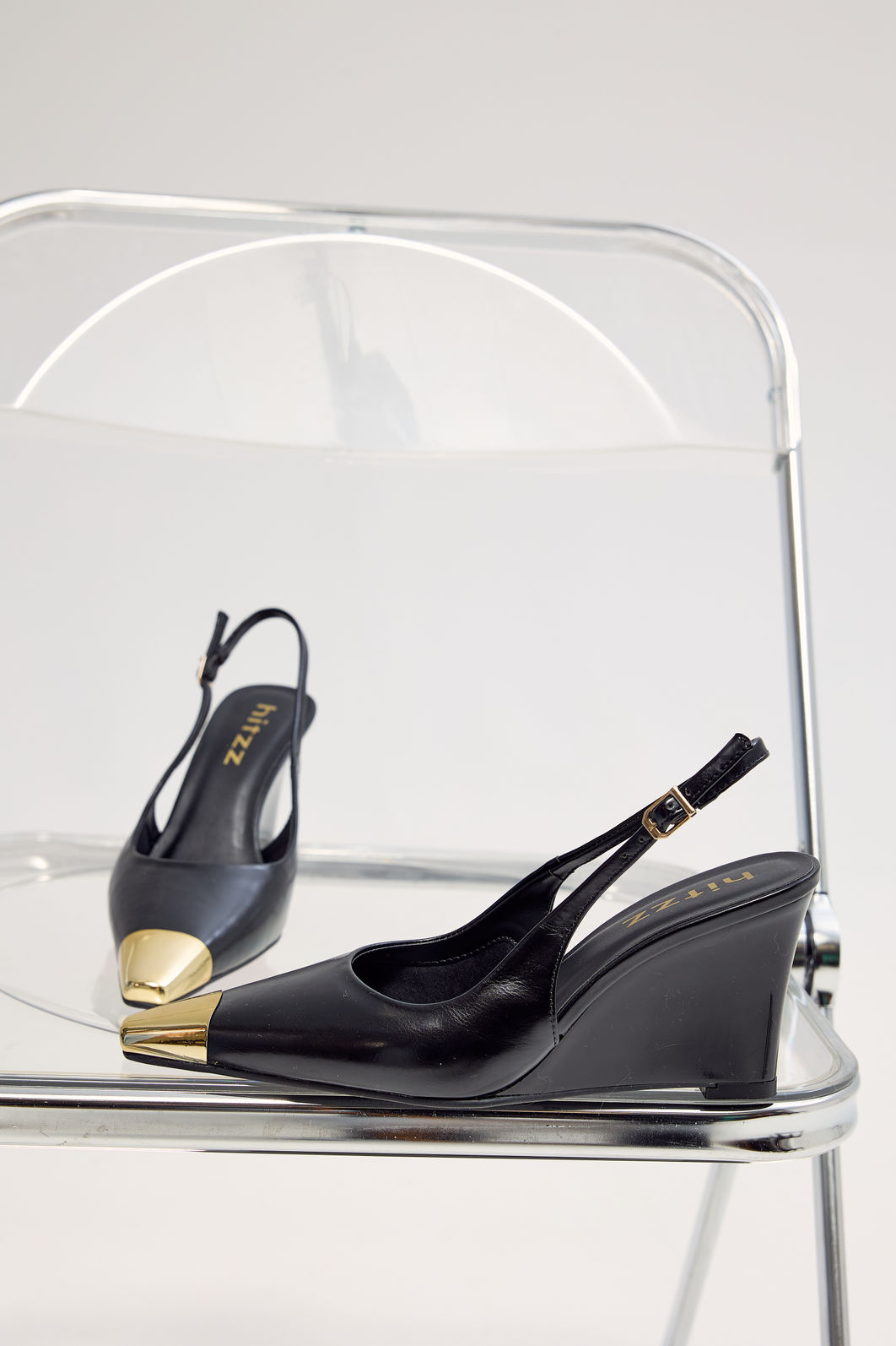 Slingback Feminino Naomi Preto Lifestyle Par de slingbacks femininos Naomi pretos com salto anabela e bico metálico dourado sobre cadeira cromada, destacando o design sofisticado do modelo.