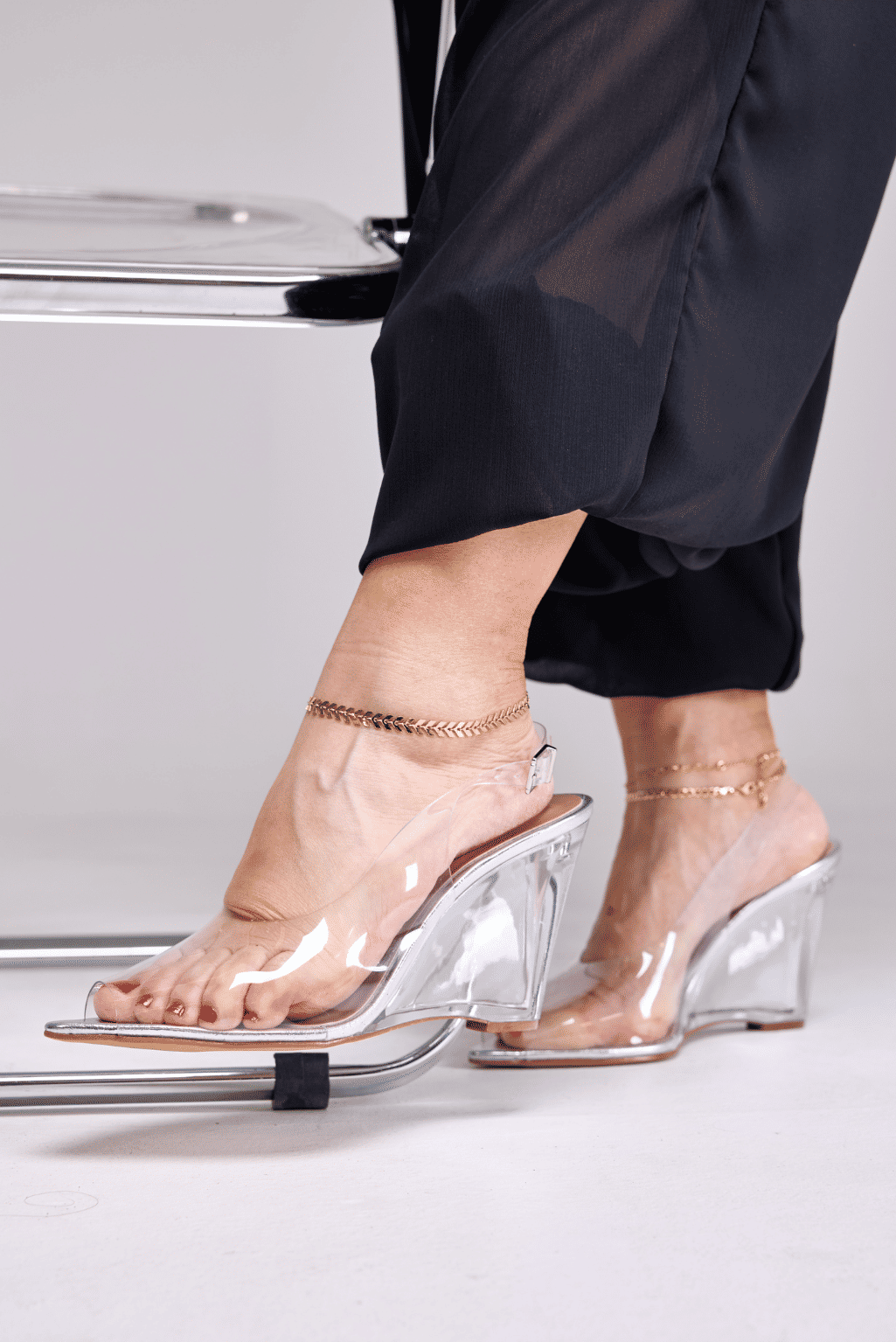 scarpin-anabela-feminina-vinil-prata-storm Detalhe dos pés com Scarpin Anabela Feminina Vinil Prata - Storm, calça preta fluida e tornozeleiras douradas, apoiado em cadeira metálica cromada