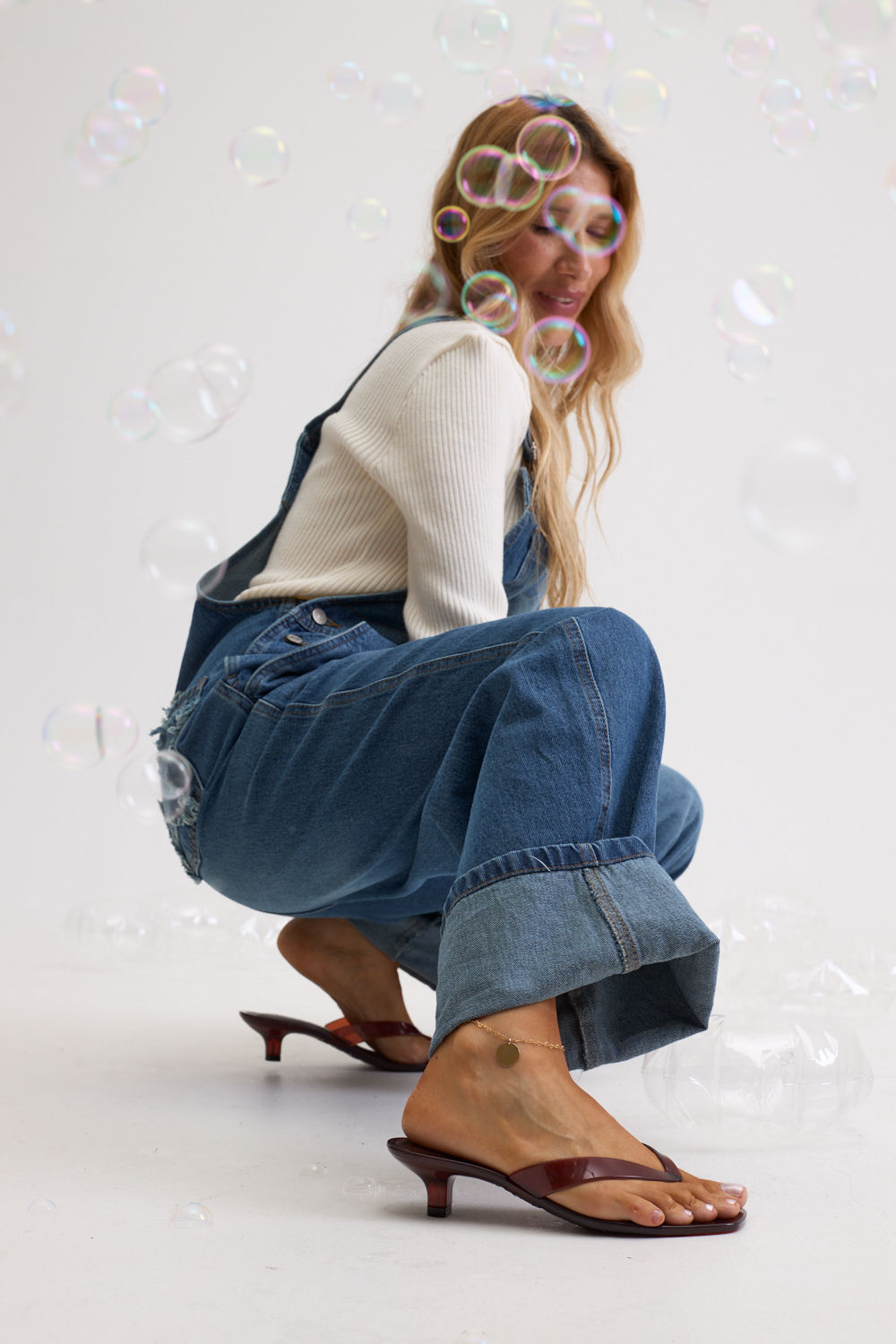 gummy-tamanco-feminino-transparente-salto-baixo-marrom Tamanco Feminino Flip Flop Gummy marrom em PVC translúcido, modelo agachada usando macacão jeans, vista lateral completa