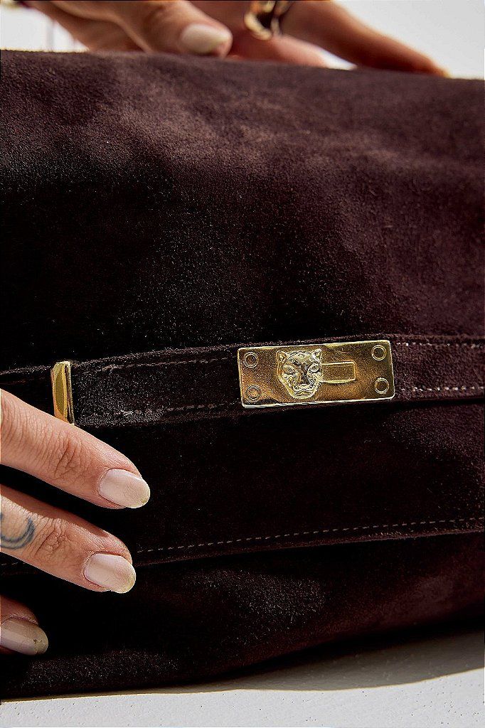 bolsa-feminina-ombro-marrom-vick Close da mão tocando o fecho dourado com detalhe de onça da bolsa de couro marrom, destacando a textura em camurça.