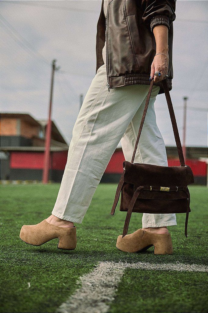 bolsa-feminina-ombro-marrom-vick Modelo carregando a bolsa de couro marrom na alça longa a tiracolo, caminhando em campo de futebol, look casual com calça branca.