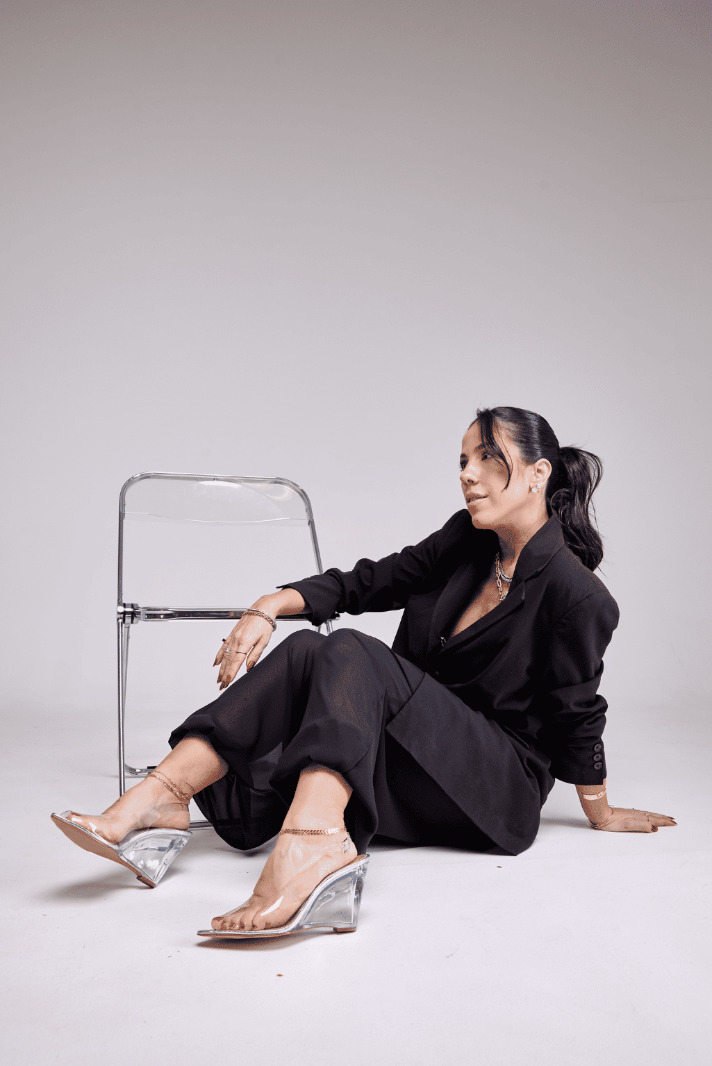 scarpin-anabela-feminina-vinil-prata-storm Scarpin Anabela Feminina Vinil Prata - Storm nos pés da modelo sentada no chão, blazer e calça preta oversized, apoiada em cadeira acrílica transparente