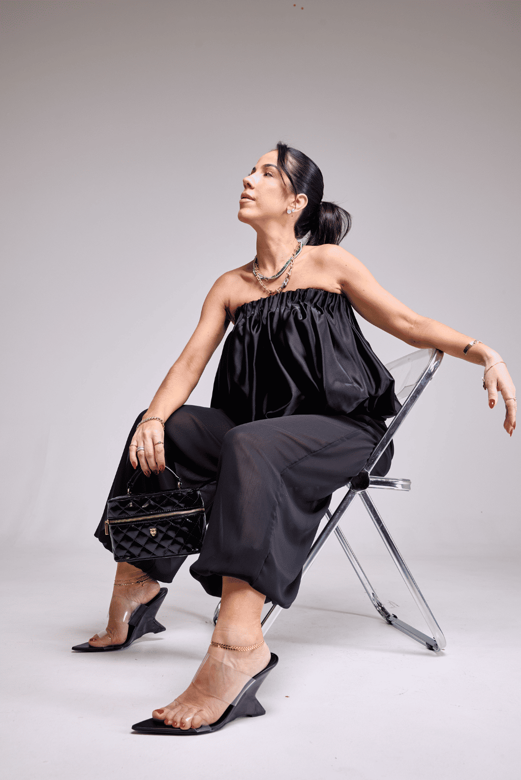 tamanco-feminino-tiras-de-vinil-preto-miranda Modelo sentada em cadeira acrílica, look total black tomara que caia, calçando o Tamanco Feminino Tiras de Vinil Preto - Miranda, corpo inteiro