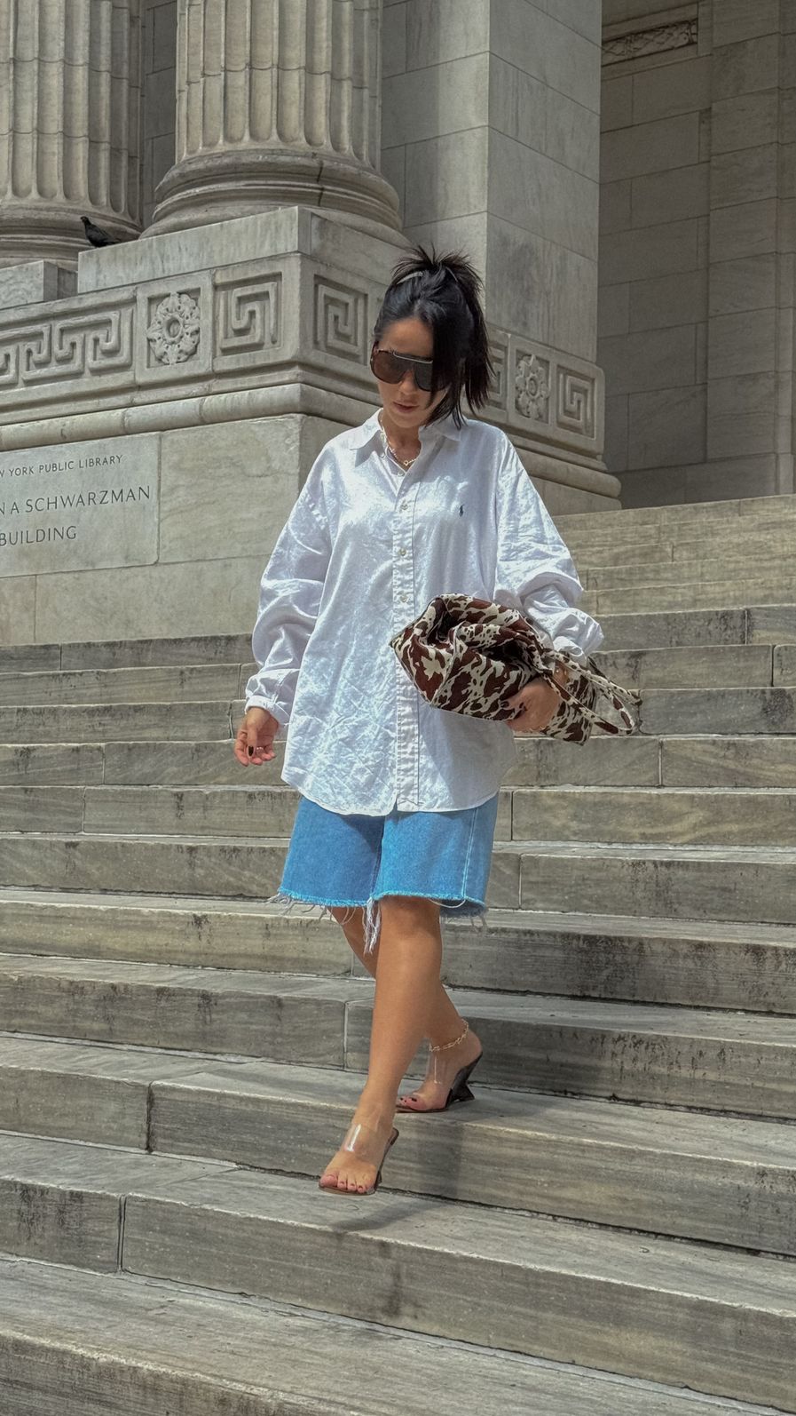 tamanco-feminino-tiras-de-vinil-marrom-miranda Modelo em look de rua descendo escadaria com camisa branca oversized e short jeans azul, usando o Tamanco Feminino Tiras de Vinil Marrom - Miranda.