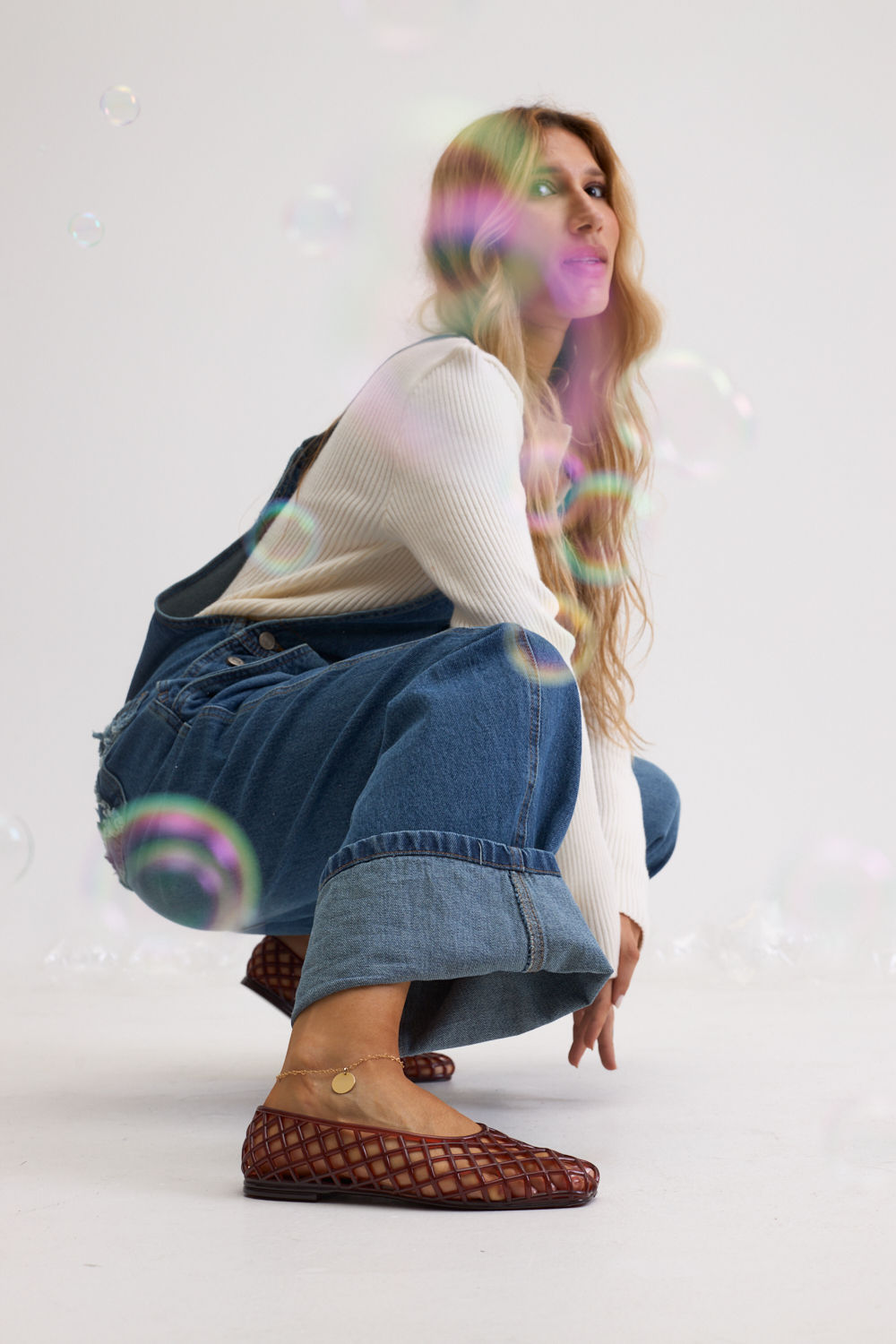 sapatilha-feminina-marrom-gummy Modelo agachada usando a Sapatilha Feminina Marrom - Gummy com tricô canelado creme e jeans wide leg índigo, composição editorial com efeito de luz e tornozeleira dourada.