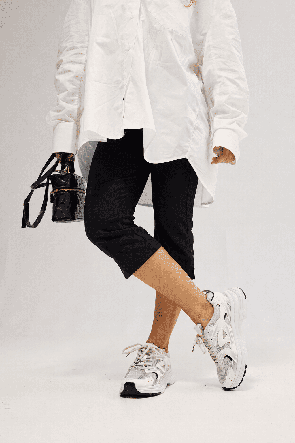 Look com Tênis Victoria Off White Modelo usando Tênis Feminino Victoria Off White com camisa branca oversized, calça preta cropped e bolsa preta, com as pernas cruzadas.