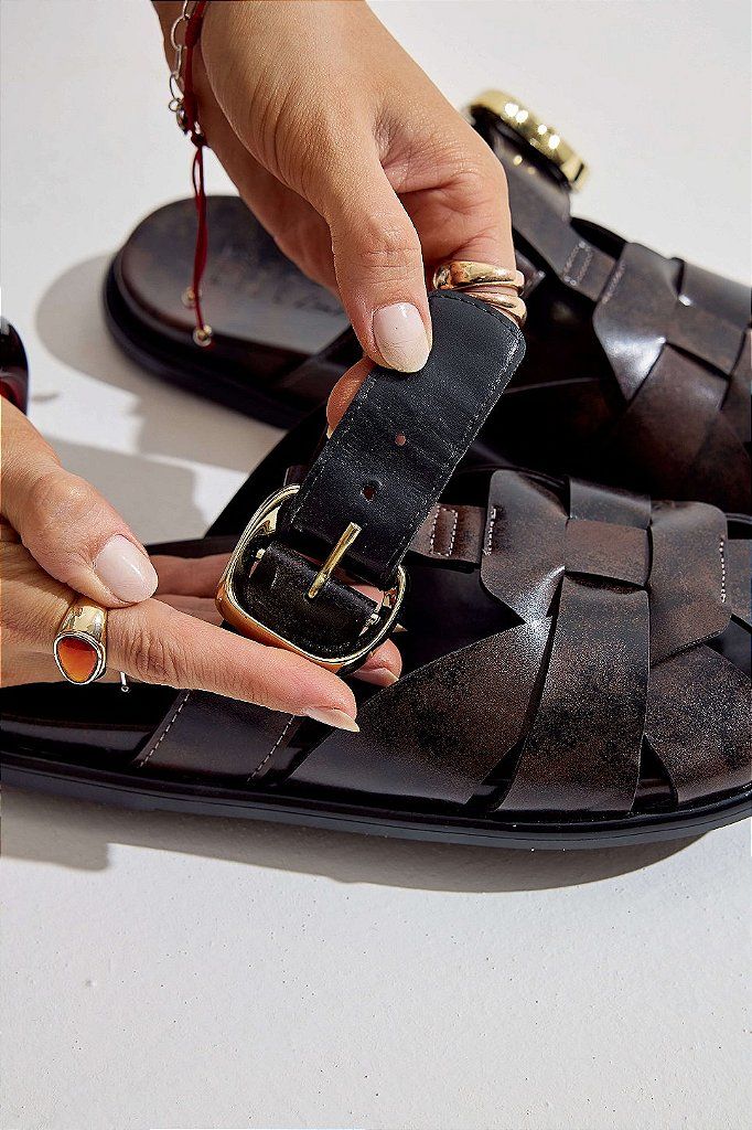 clog-feminino-fivela-marrom-aura Mãos ajustando a fivela dourada do clog marrom, demonstrando os furos de regulagem da tira.