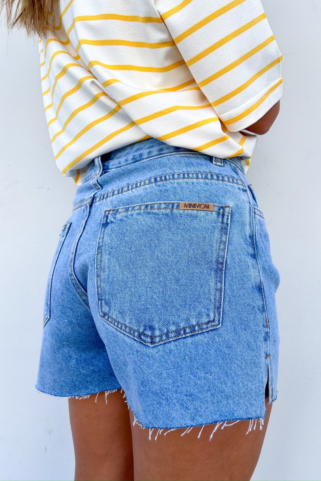 Detalhe da Abertura Lateral em Shorts Jeans Close-up da lateral do Shorts Domênica, destacando a pequena abertura na perna e a textura do jeans 100% algodão.