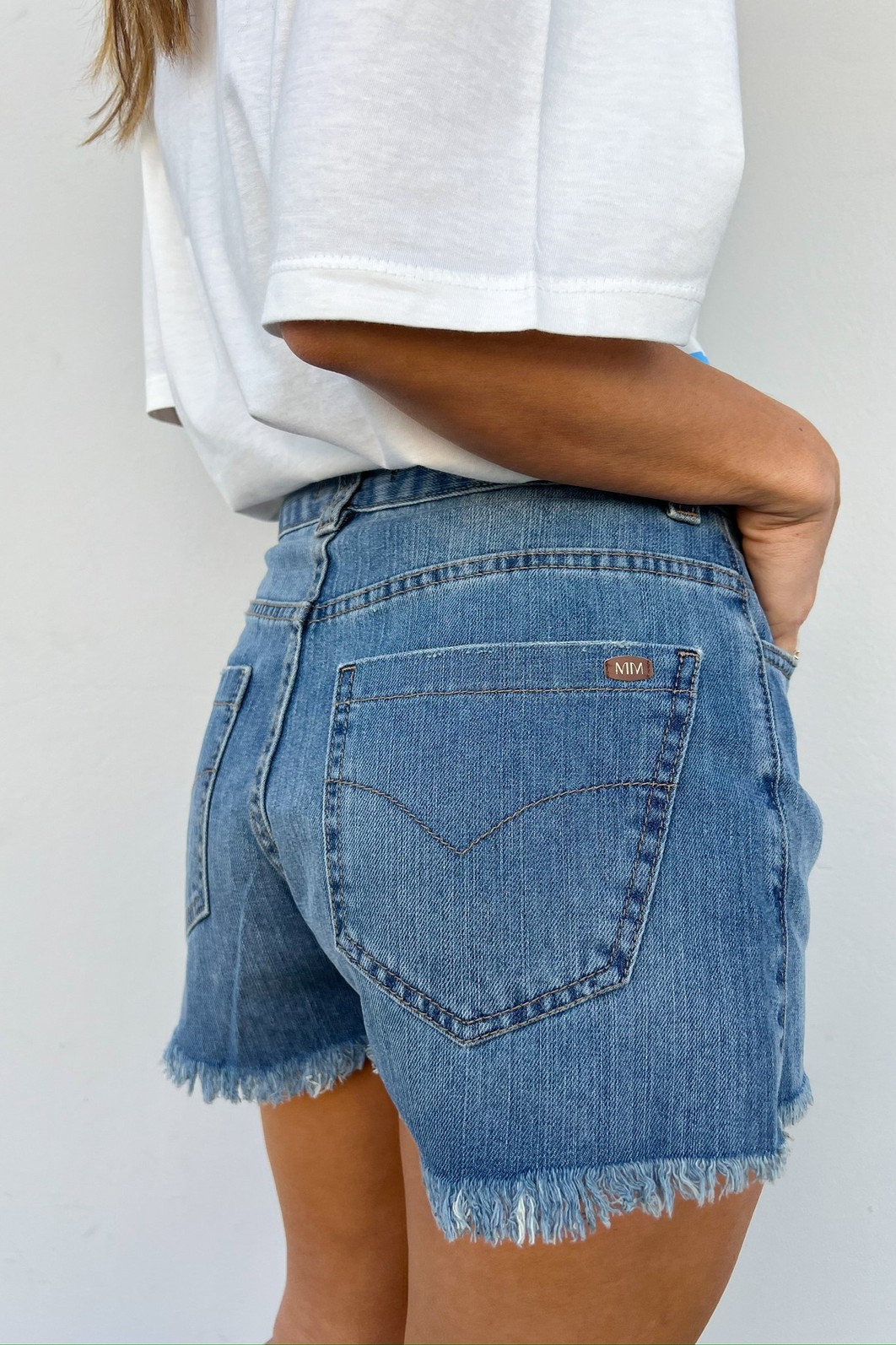Shorts Jeans Denim Cleo – Detalhes das costas com bolsos e acabamento desfiado Parte traseira do shorts jeans feminino azul, com bolsos aplicados, acabamento moderno e barra desfiada, peça básica e versátil da MiniMoni.