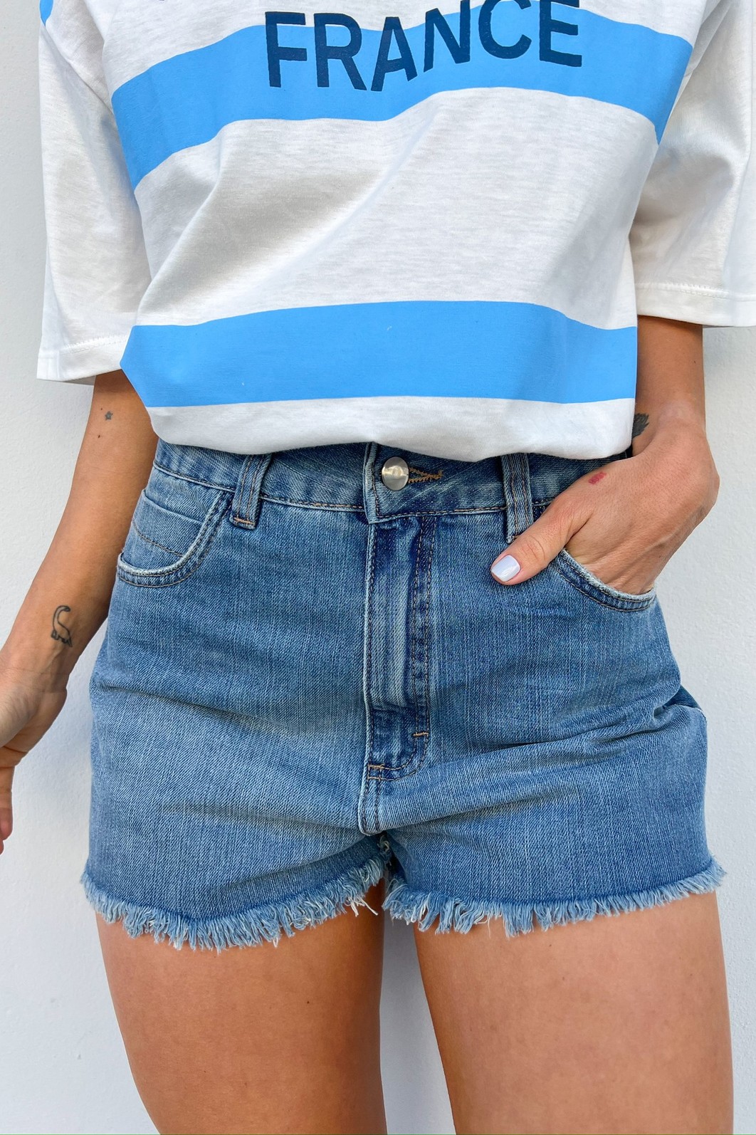 Shorts Jeans Denim Cleo – Look casual feminino com blusa branca Look casual feminino composto por shorts jeans azul Denim Cleo com barra desfiada e blusa branca básica, ideal para dia a dia e passeios.