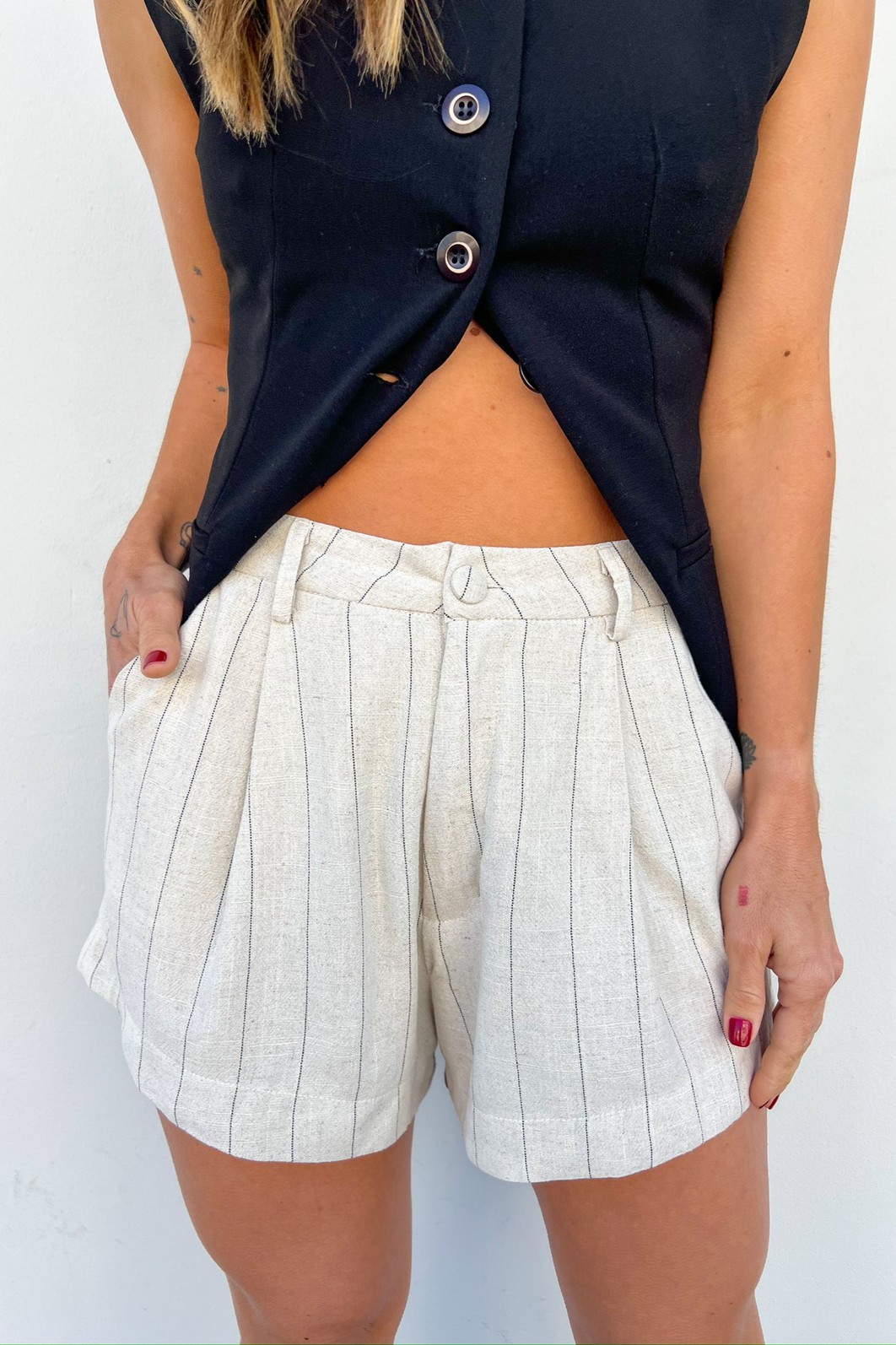 Look com shorts de alfaiataria e colete de alfaiataria preto. Modelo usando o Shorts de alfaiataria com risca de giz e um colete com uma proposta aberta, mostrando os detalhes do shorts, seu botão forrado e também a versatilidade da peça.