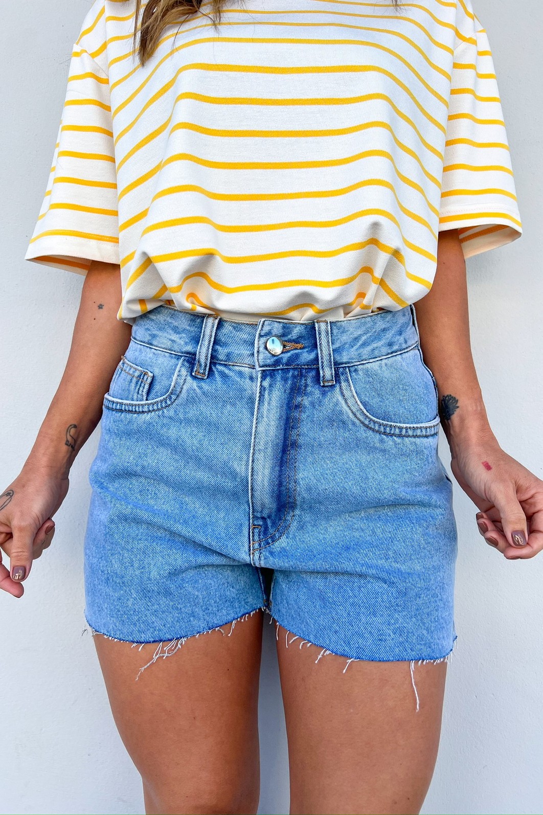 Shorts Jeans Feminino com Bainha Desfiada e Corte Reto Modelo vestindo o Shorts Jeans Domênica, mostrando a modelagem reta, a lavagem clássica e o detalhe da bainha desfiada.