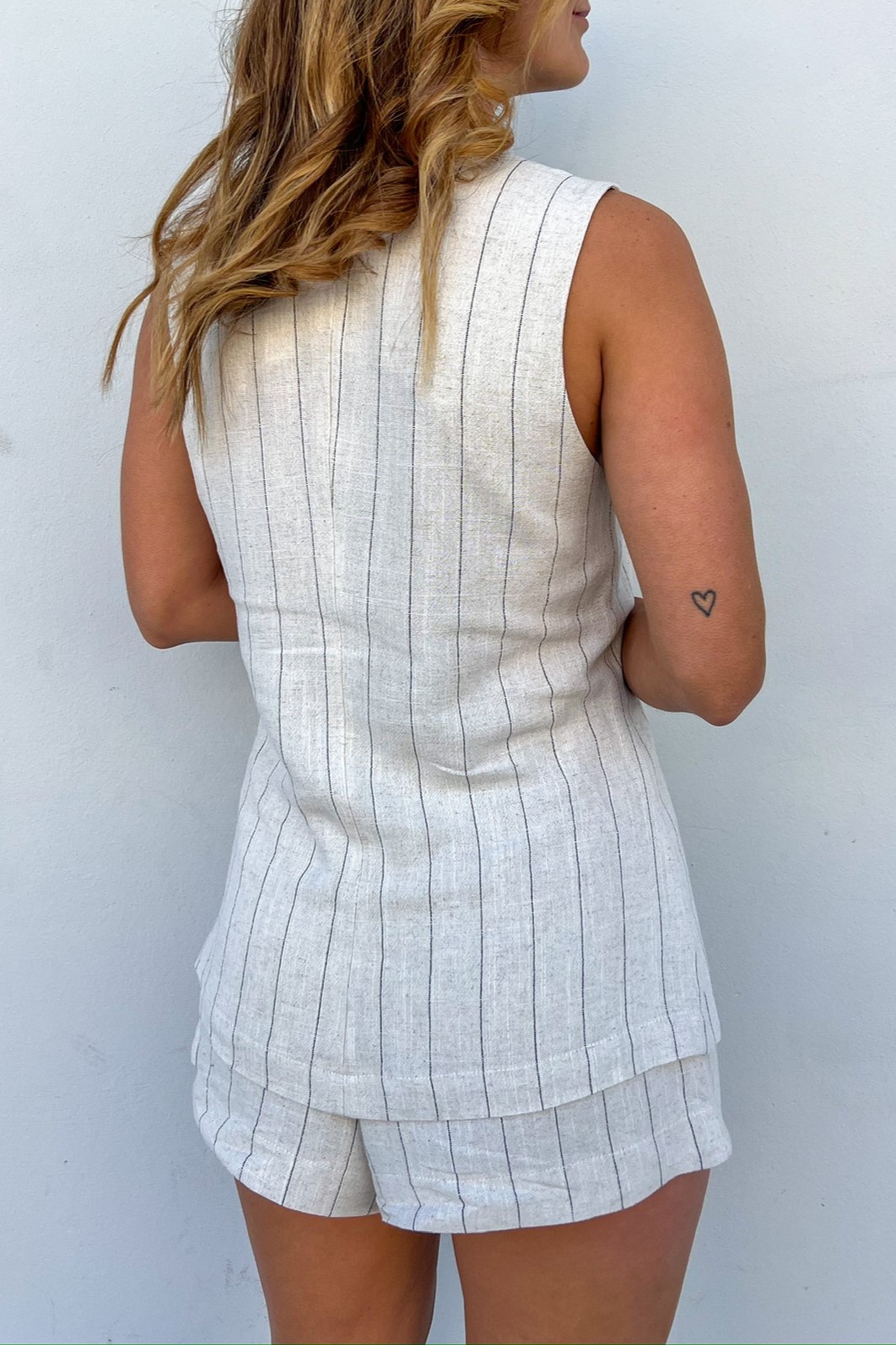 Conjunto de alfaiataria risca de giz mostrado de costas. Modelo de costas mostrando o caimento do colete de alfaiataria combinado com o shorts de alfaiataria risca de giz, mostrando um look leve e confortável.