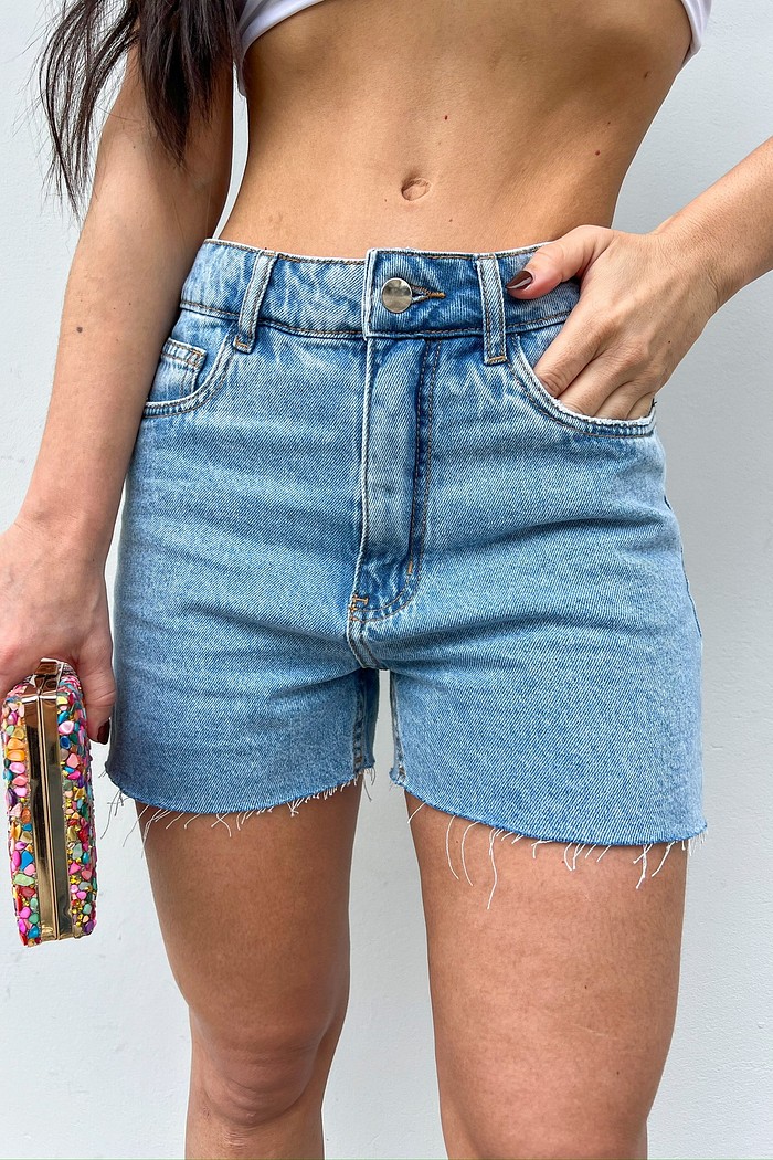 Shorts Jeans Bia — vista frontal Shorts Jeans Bia MiniMoni em denim azul-claro, barra desfiada, vista frontal com mão no bolso, foco no caimento
