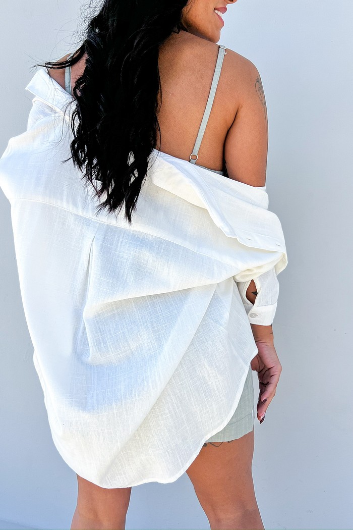 Camisa oversized branca – estilo casual elegante Camisa feminina oversized branca de manga longa em tecido leve, usada sobre alça fina cinza, look casual elegante visto de costas.