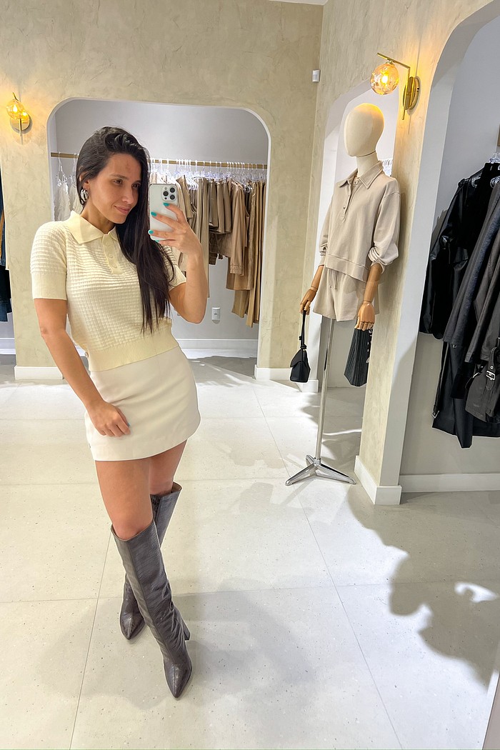 Look elegante com tricô bege e saia branca Look feminino com blusa de tricô bege de manga curta e gola polo, combinada com saia branca curta e bota cano alto marrom.