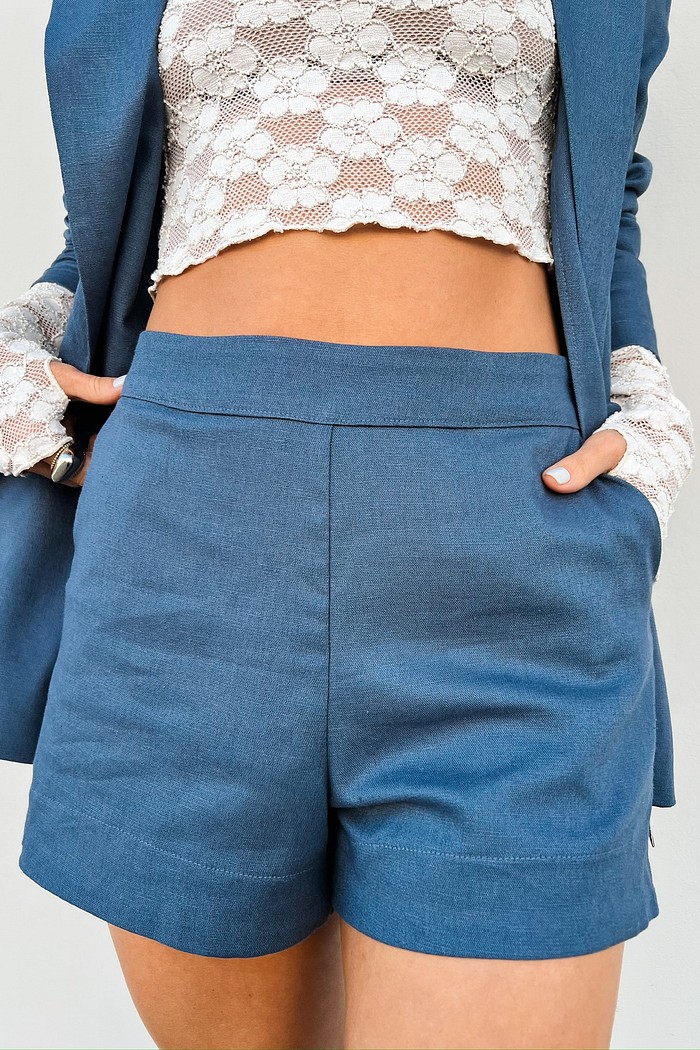 Shorts Alfaiataria Azul Jeans com Cós Largo Estruturado Vista de frente de uma mulher usando o Shorts de Alfaiataria Azul Sylvie, destacando o corte impecável na cintura, o cós largo sem botões e a textura sutil do tecido com linho.