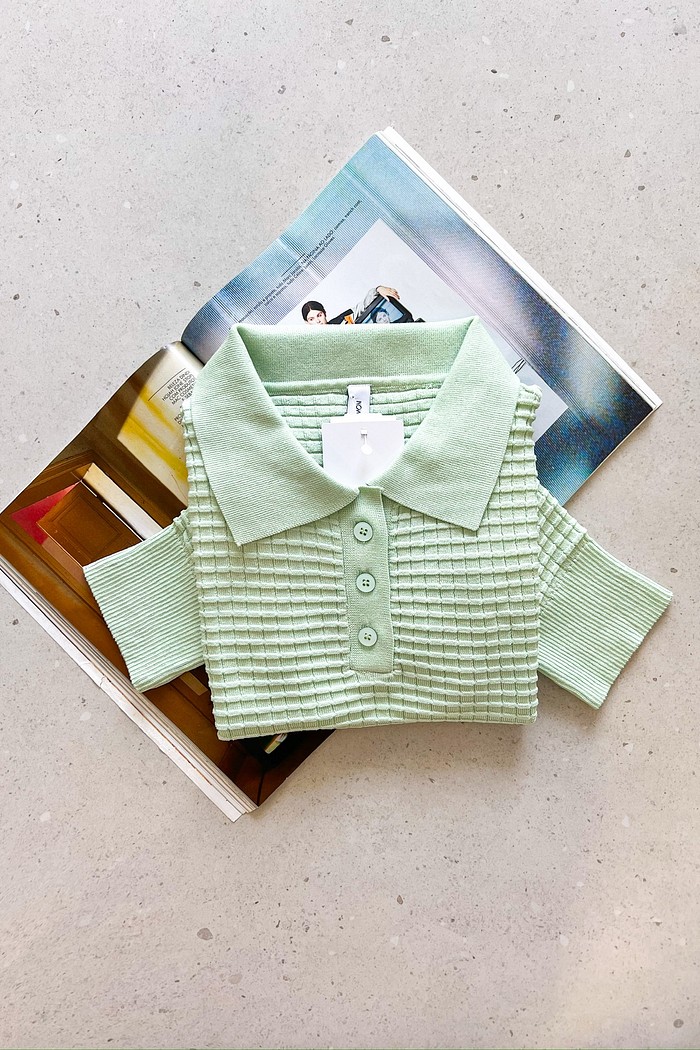 Blusa polo verde clara em tricô texturizado Blusa polo feminina de tricô verde claro, manga curta, com textura quadriculada e botões frontais, dobrada sobre revista.