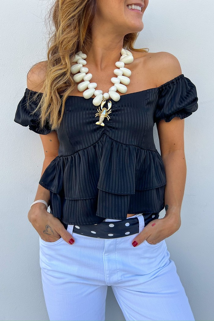 Mulher usando blusa preta ombro a ombro com babados e mangas curtas bufantes, combinada com calça branca.