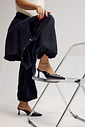 Mule Zara Preto Salto Taça Modelo com um dos pés apoiado em cadeira transparente usando mule feminino Zara preto de bico fino, salto taça e aplicações metálicas.