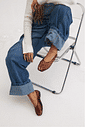 Modelo sentada em cadeira acrílica com estrutura metálica, calça jeans wide leg e blusa de tricô creme, exibindo os pés calçados com a Sapatilha Feminina Marrom - Gummy vazada e tornozeleiras douradas