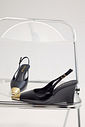 Slingback Feminino Naomi Preto Lifestyle Par de slingbacks femininos Naomi pretos com salto anabela e bico metálico dourado sobre cadeira cromada, destacando o design sofisticado do modelo.