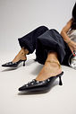 Mule Feminino Preto com Aplicações Modelo sentada no chão usando mule feminino preto Zara com aplicações metálicas prateadas, evidenciando o design elegante e o salto taça.