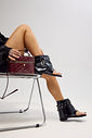 eva-sandalia-feminina-rasteira-fivelas-preta Modelo sentada usando a Bota Sandália Eva Preta feminina com bico quadrado, solado rasteiro, tira para o dedo e duas fivelas metálicas ajustáveis, combinada com bolsa vinho matelassê.