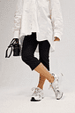 Look com Tênis Victoria Off White Modelo usando Tênis Feminino Victoria Off White com camisa branca oversized, calça preta cropped e bolsa preta, com as pernas cruzadas.