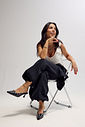Mule Zara Preto nos Pés Modelo sentada usando mule feminino Zara preto com bico fino, salto taça e aplicações metálicas prateadas, combinado com calça preta ampla e regata branca.