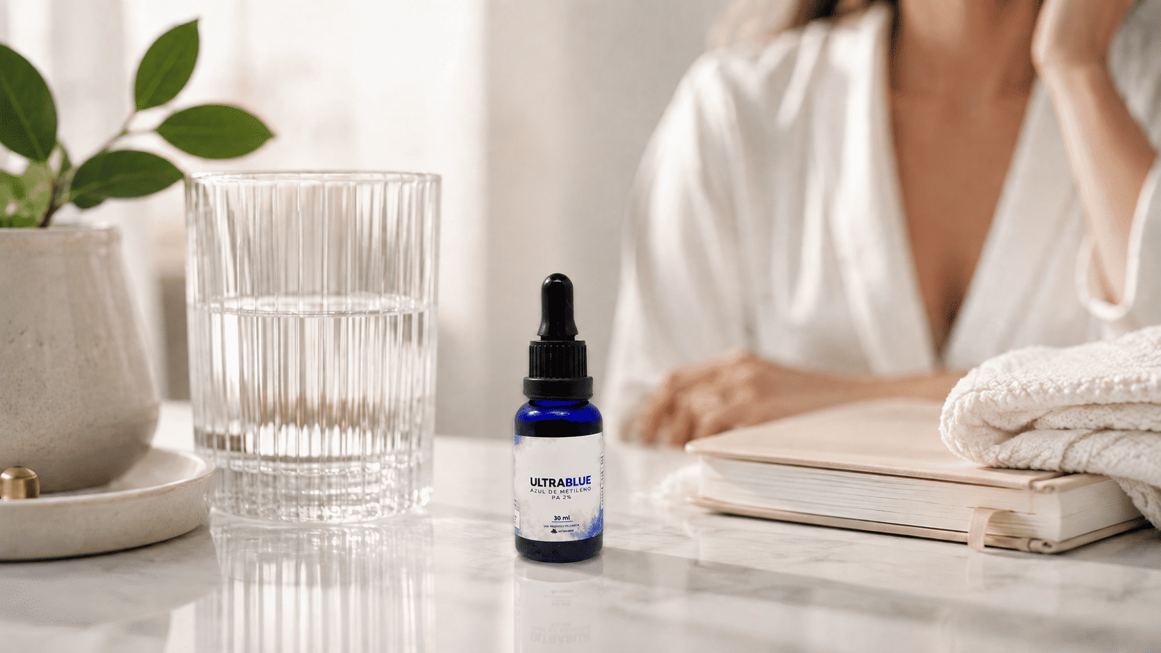 Frasco azul com conta-gotas do produto UltraBlue Azul de Metileno PA 2% em 30ml sobre uma superfície de mármore branco, ao lado de um copo de água transparente. Ao fundo, uma mulher de robe branco e um caderno empilhado com toalhas compõem um ambiente clean e minimalista.