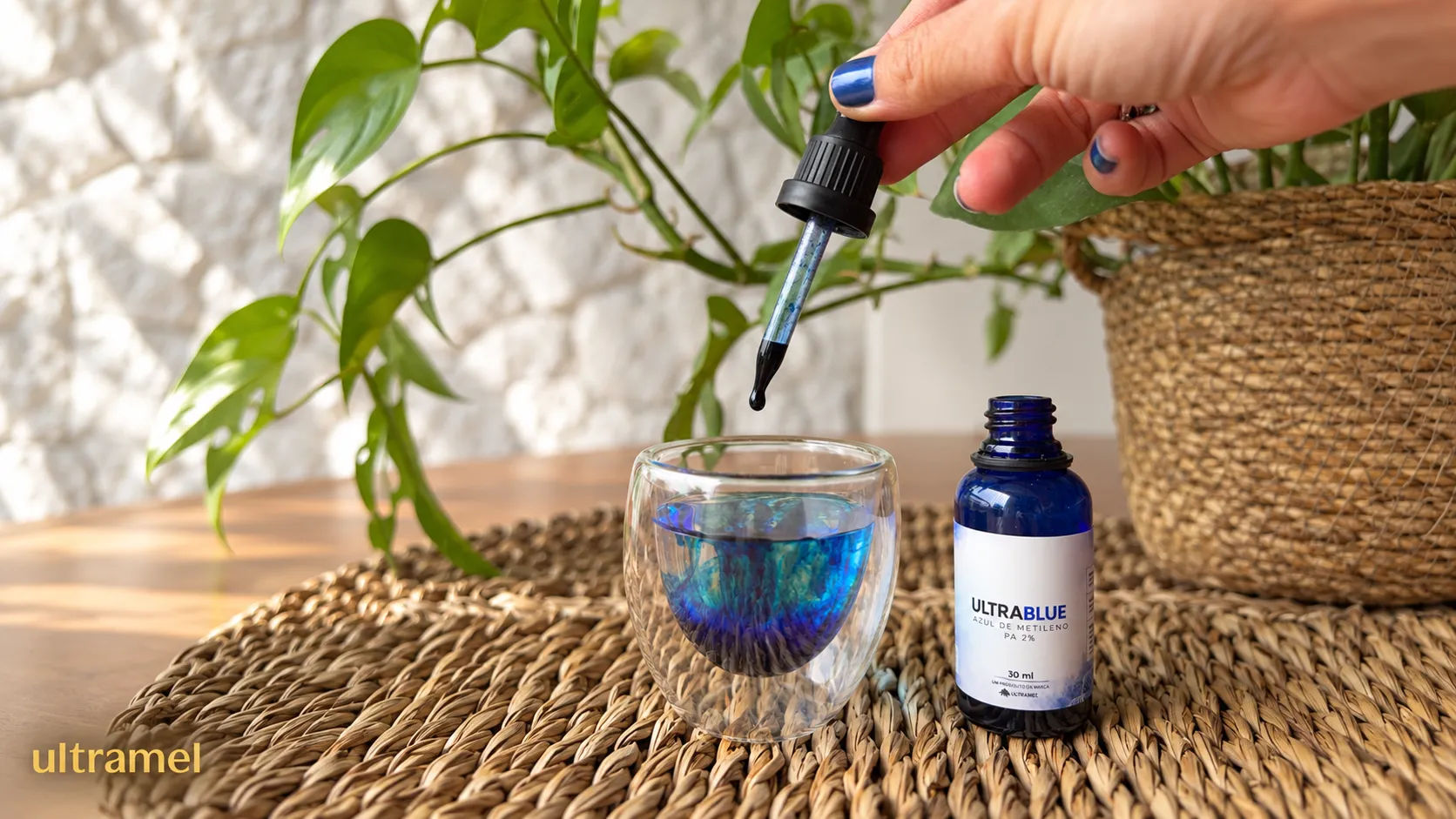 Mão segurando um conta-gotas com Azul de Metileno (produto UltraBlue PA 2% da Ultramel) sendo pingado em um copo de vidro com água, tingindo-a de azul, ao lado do frasco original de 30ml sobre uma superfície de palha.