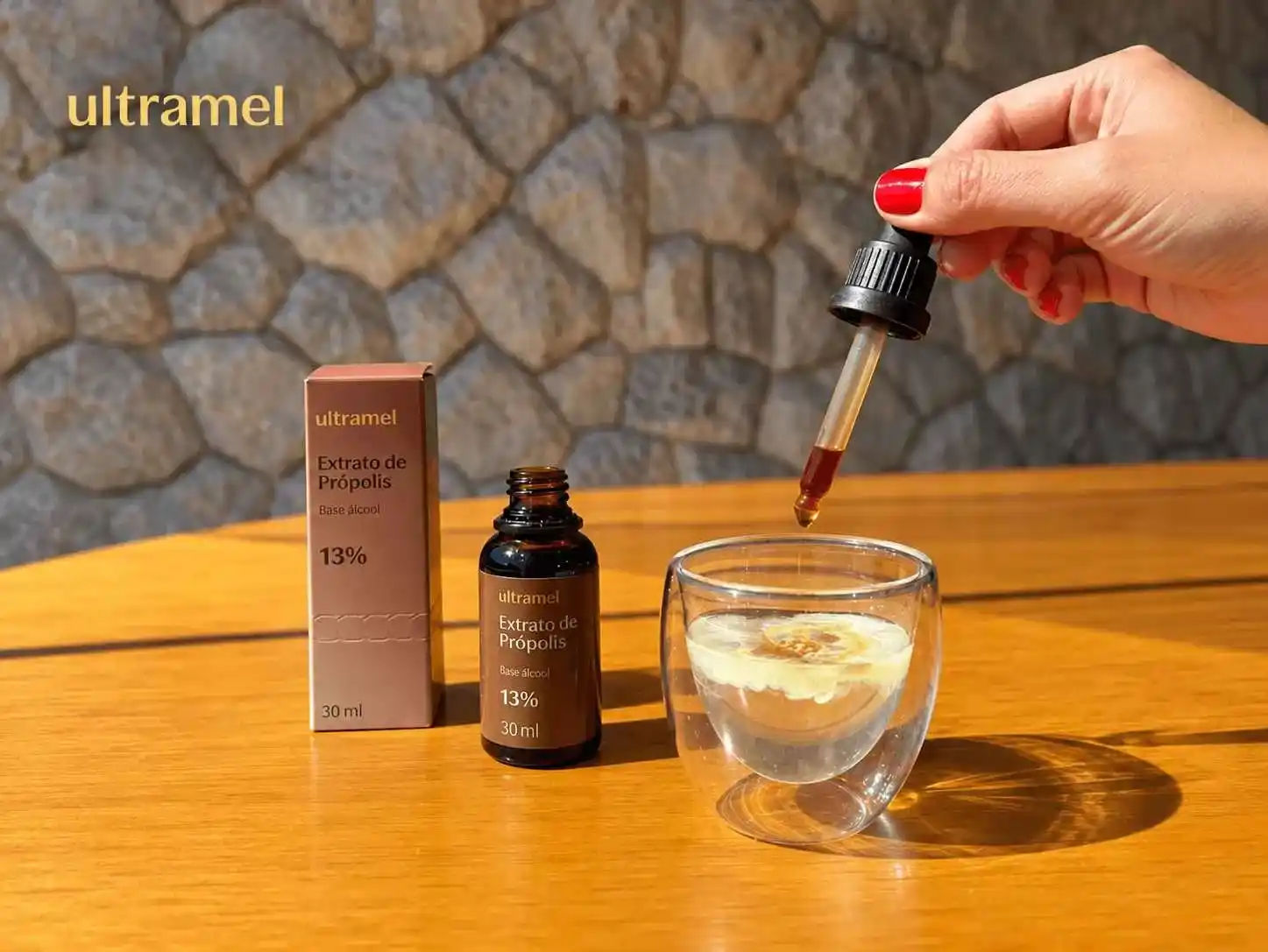 Mão segurando um conta-gotas com extrato de própolis da marca Ultramel sobre um copo de chá, ao lado da embalagem do produto Extrato de Própolis Base Álcool 13% 30ml. O frasco âmbar e a caixa rosa estão apoiados sobre uma mesa de madeira.