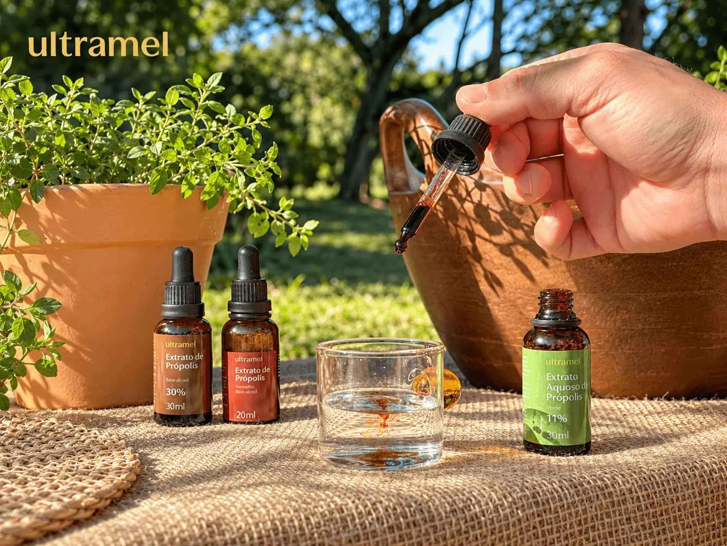 Mão segurando um conta-gotas com extrato de própolis sendo adicionado a um copo com água, ao lado de três frascos da marca Ultramel: Extrato de Própolis 30% (30ml), Extrato de Própolis Vermelho (20ml) e Extrato Aquoso de Própolis Verde 11% (30ml). Os produtos estão dispostos sobre uma superfície de juta, em ambiente externo com plantas ao fundo.