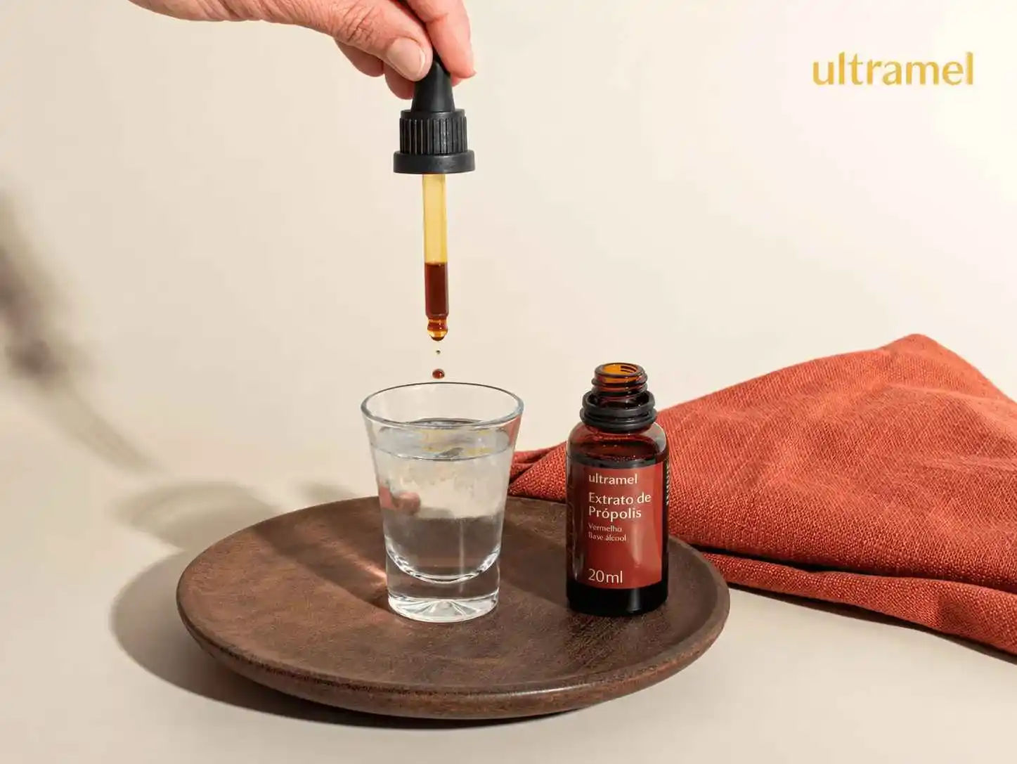 Mão segurando um conta-gotas com extrato de própolis vermelho Ultramel, pingando gotas em um copo com água. Ao lado, o frasco de 20ml do Extrato de Própolis Vermelho Base Álcool da Ultramel está apoiado em um prato de madeira.
