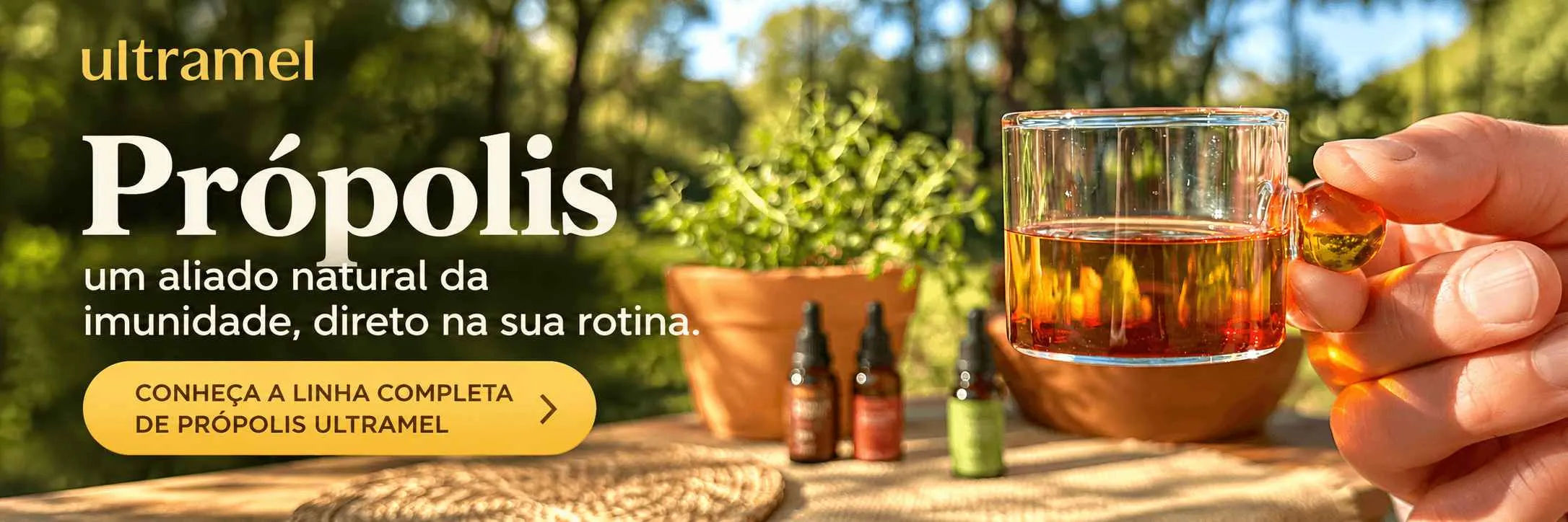 Mão segurando um copo de vidro com extrato de própolis diluído em água, ao lado de frascos de própolis em gotas da marca Ultramamel sobre uma mesa ao ar livre.