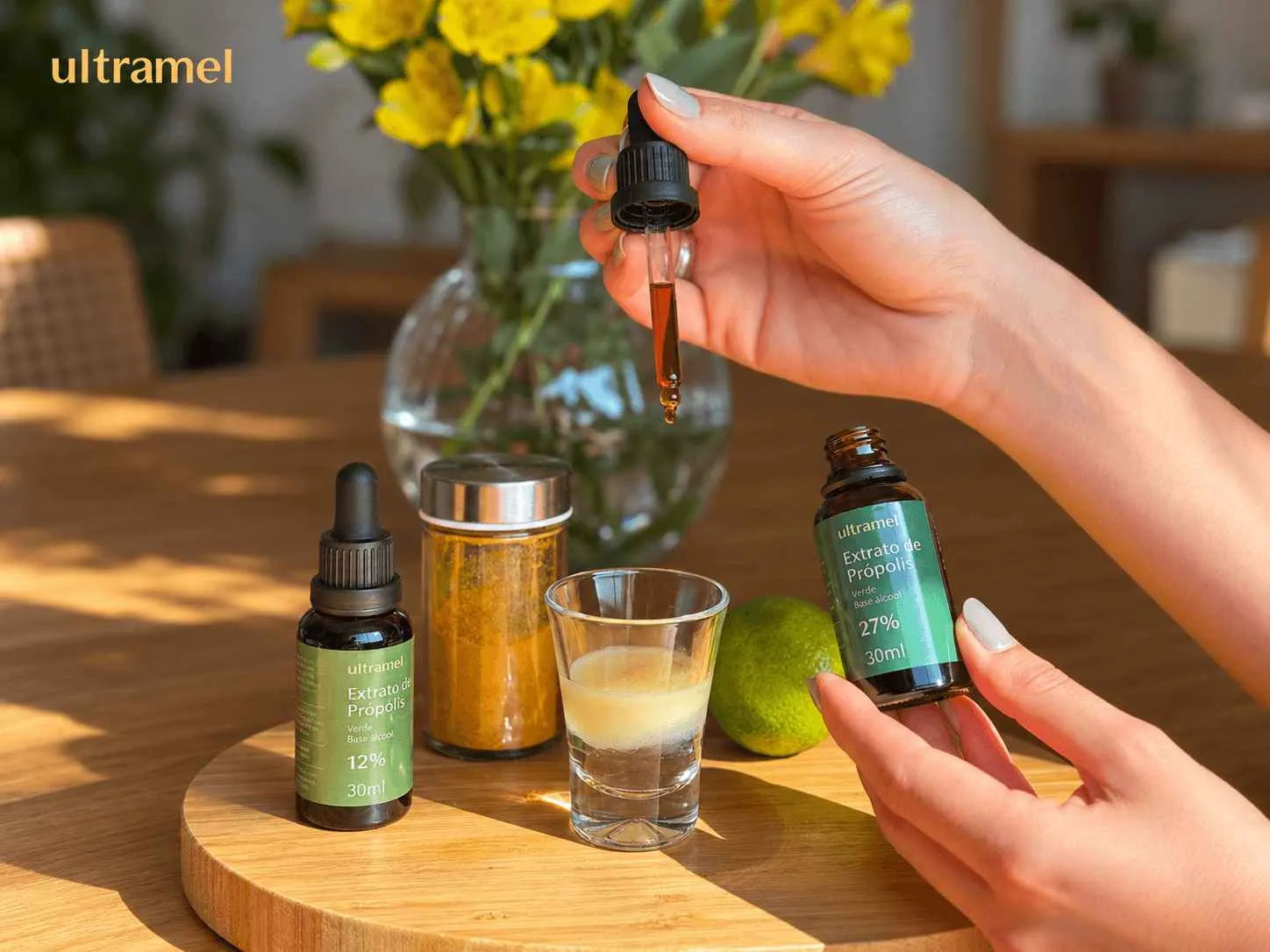 Mãos segurando um frasco gotejador e uma embalagem de Extrato de Própolis Verde Ultramel 27% 30ml, ao lado de outro frasco da versão 12%, um shot com líquido e uma lima sobre uma bandeja de madeira. A cena ilustra o uso e comparação de diferentes concentrações de extrato de própolis verde em base álcool.