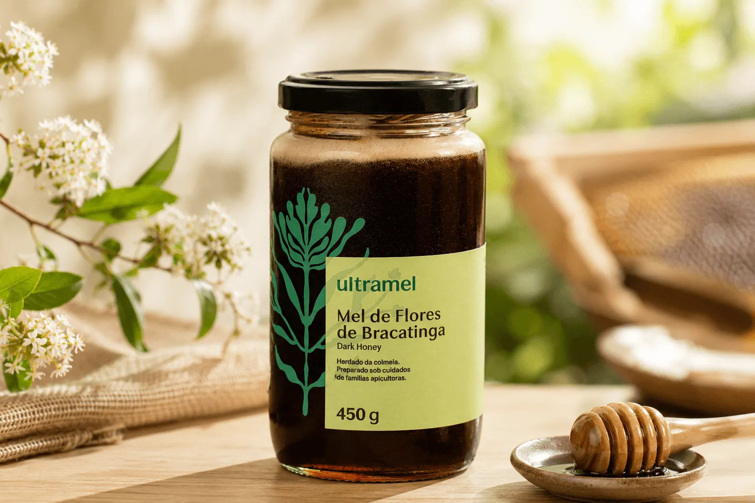 Pote de vidro de 450g de Mel de Flores de Bracatinga Dark Honey da marca Ultramel sobre uma mesa de madeira, acompanhado de uma colher de mel e flores brancas ao fundo. O mel escuro é visível através do vidro transparente, destacando a cor característica desse tipo de mel.