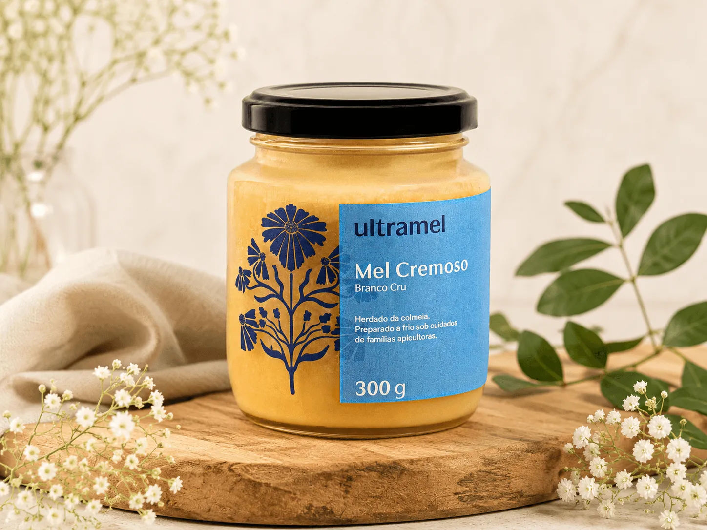 Pote de vidro de Mel Cremoso Branco Cru da marca Ultramel, com 300g, sobre uma tábua de madeira rústica decorada com flores brancas delicadas. O rótulo azul destaca informações sobre o produto, que é preparado a frio sob cuidados de famílias apicultoras.