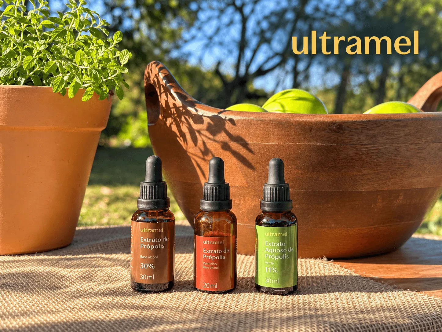 Três frascos de extrato de própolis da marca Ultramel — nas versões base álcool 30ml, vermelho base álcool 20ml e aquoso verde 11% 30ml — dispostos sobre uma superfície de juta, com um vaso de plantas e uma tigela de madeira com frutas ao fundo.
